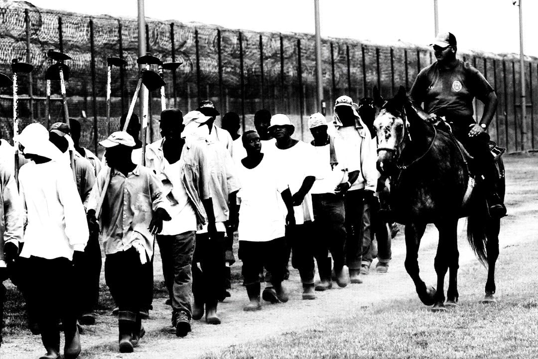 A group of prison laborers walking in single file, accompanied by a mounted guard, in a monochrome correctional facility setting. The fence is visible in the background.