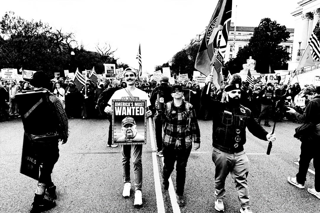 A group of protesters marching on a street, holding signs and flags. One person in the foreground carries a sign reading "America's Most Wanted" with Donald Trump's ugly face below, while others wear various clothing styles. The scene is black and white.